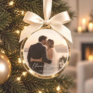 Pode incluir: Um ornamento redondo de vidro com borda dourada, apresentando uma fotografia impressa de um casal abraçado ao pôr do sol. O ornamento é finalizado com um laço de cetim creme e exibe o texto branco Olivia & James e 10.10.2026.
