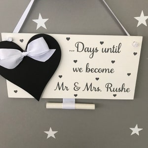May include: White wooden sign with a black heart and white ribbon. The sign says "... Days until we become Mr & Mrs. Rushe".