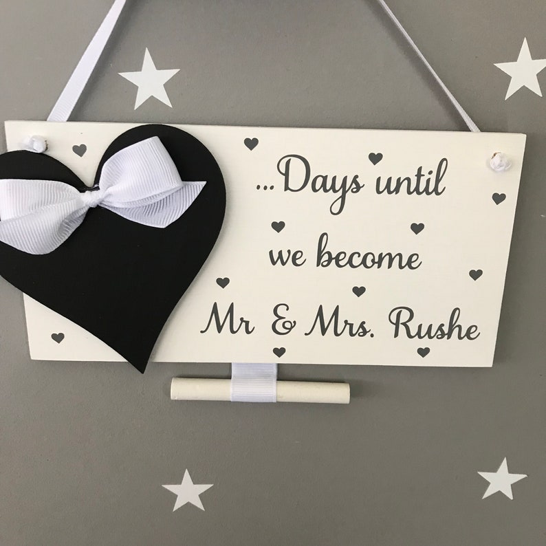 May include: White wooden sign with a black heart and white ribbon. The sign says "... Days until we become Mr & Mrs. Rushe".