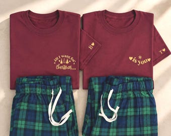 May include: Two maroon short-sleeved t-shirts and two pairs of green and blue plaid pajama pants. The left shirt features gold text reading "all I want for Christmas..." with tree icons. The right shirt features gold text "is you" with hearts.