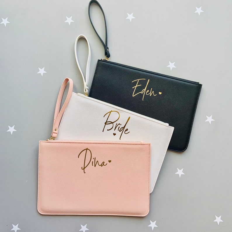Peut inclure: Trois pochettes avec des lettres dor&eacute;es. La pochette du haut est noire avec le nom "Eden" &eacute;crit en or. La pochette du milieu est blanche avec le mot "Bride" &eacute;crit en or. La pochette du bas est rose avec le nom "Diha" &eacute;crit en or.