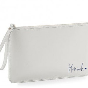May include: A white clutch purse with a zipper closure and a wrist strap. The purse has the name "Hannah" written in blue cursive on the front with a small heart.