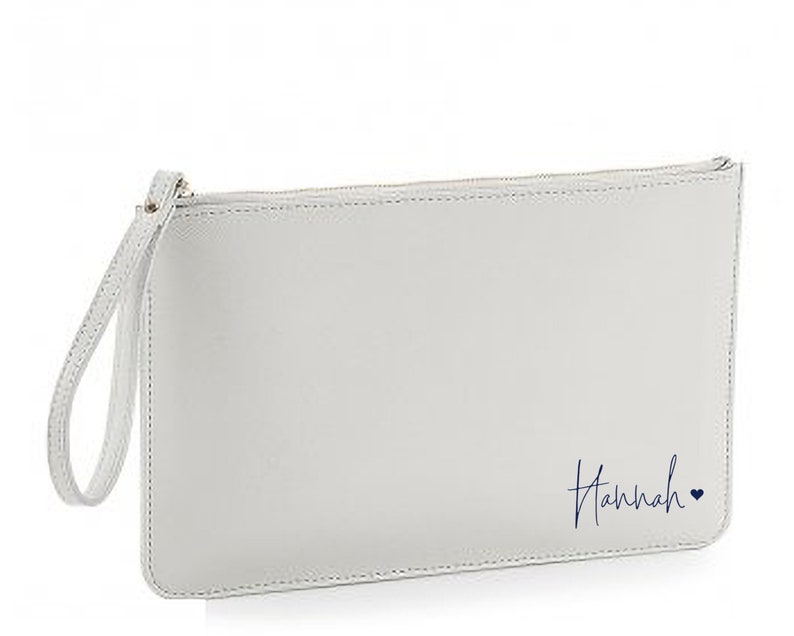 May include: A white clutch purse with a zipper closure and a wrist strap. The purse has the name "Hannah" written in blue cursive on the front with a small heart.