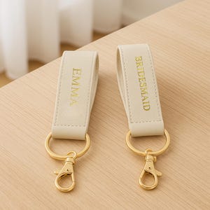 May include: Two cream-coloured keyrings with gold-coloured metalwork. One keyring has "EMMA" in gold lettering, the other reads "BRIDESMAID". They are on a light wood surface.