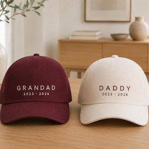 Peut inclure: Deux casquettes de baseball, une bordeaux et une crème. La casquette bordeaux porte l'inscription "GRANDAD" avec les années "2023 2026". La casquette crème dit "DADDY" avec les années "2023 2026". Elles sont sur une surface en bois.