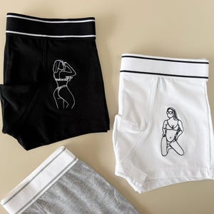 May include: Two pairs of underwear with a white graphic print of a woman in a bikini. One pair is black with a white waistband and the other is white with a black waistband.