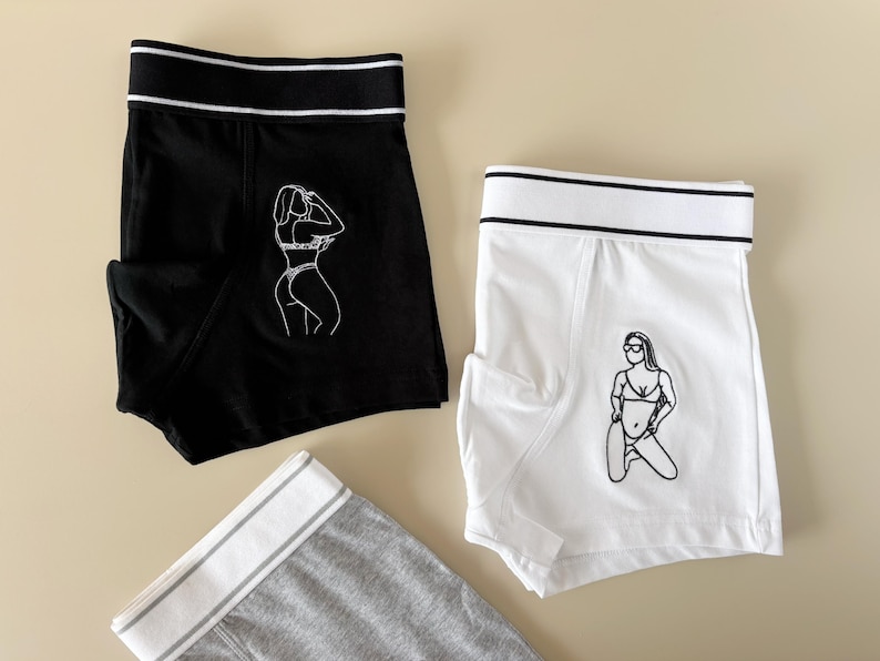 May include: Two pairs of underwear with a white graphic print of a woman in a bikini. One pair is black with a white waistband and the other is white with a black waistband.