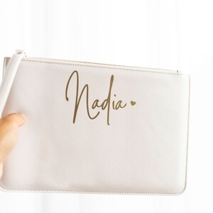 May include: A white zippered pouch with a gold inscription that reads "Nadia" with a small heart.