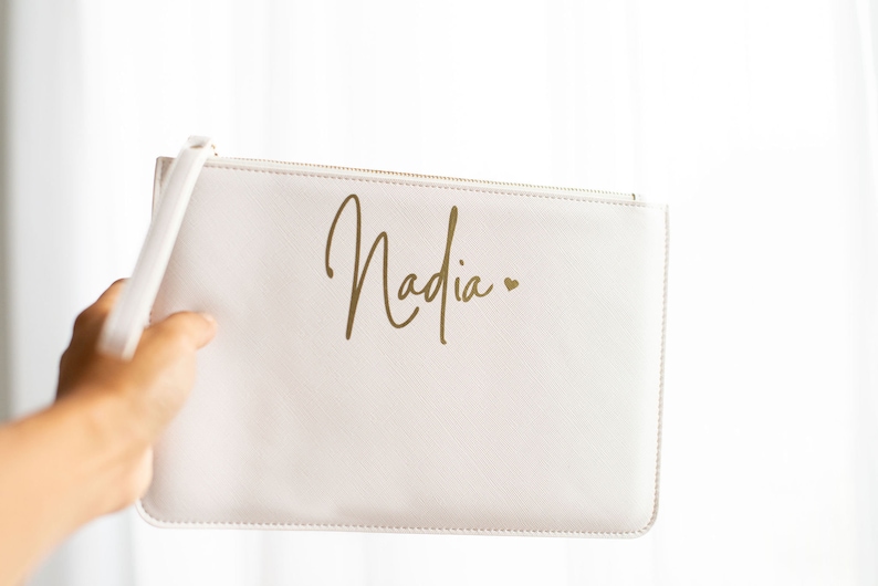 May include: A white zippered pouch with a gold inscription that reads "Nadia" with a small heart.