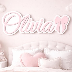 Op de afbeelding: Een roze en wit houten bord met de naam "Olivia" in sierlijke letters, met een strik. Het bord hangt aan een lichtroze muur boven een bed met witte kussens en een roze hartvormig kussen, ideaal voor een kinderkamer.