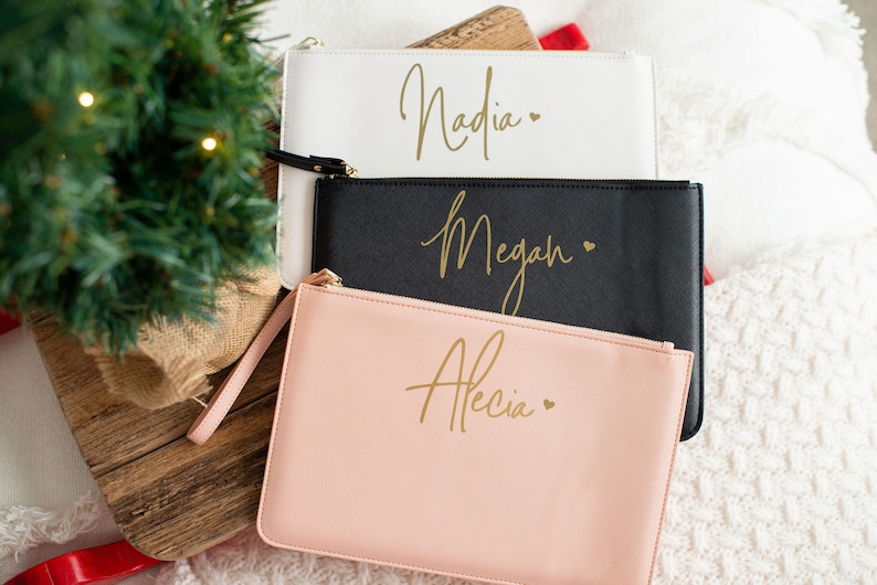 May include: Three personalized cosmetic bags with gold lettering. The bags are white, black, and pink. The names on the bags are Nadia, Megan, and Alecia.
