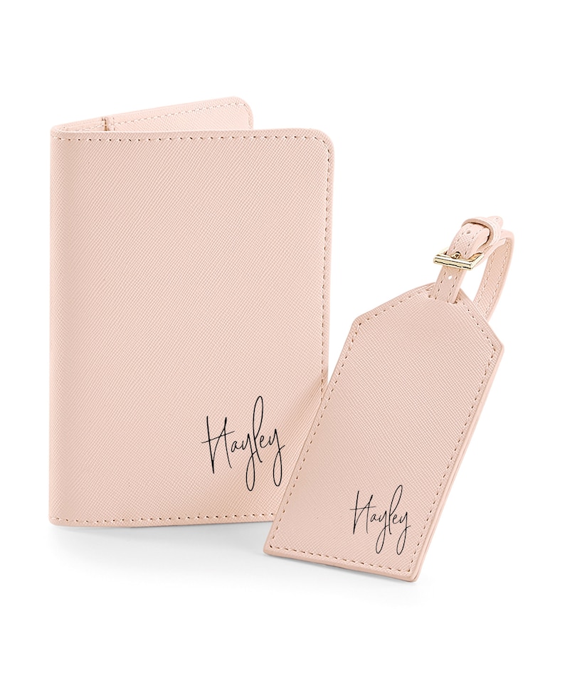 以下が含まれることがあります： 淡いピンクのパスポートホルダーと荷物タグのセット。両方に「Hayley」という名前が筆記体で書かれています。