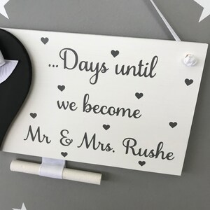 May include: White wooden sign with the text "... Days until we become Mr & Mrs. Rushe" in grey lettering. The sign has a small chalkboard attached to the bottom and is decorated with small grey hearts.