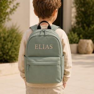 Puede incluir: Una mochila de color verde salvia con asa superior, correas ajustables para los hombros, un compartimento principal con cremallera y un bolsillo frontal con cremallera. El nombre ELIAS está bordado en hilo beige claro en el frente.