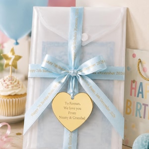 May include: A translucent white rectangular envelope secured with a light blue ribbon printed with "Happy 7th Birthday Roman, 14th December 2026". A gold heart-shaped tag hangs from the ribbon, engraved with "To Roman, We love you From Nanny & Grandad".