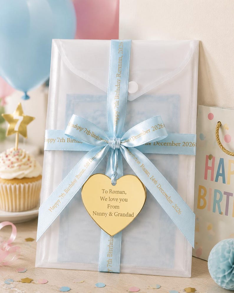 May include: A translucent white rectangular envelope secured with a light blue ribbon printed with "Happy 7th Birthday Roman, 14th December 2026". A gold heart-shaped tag hangs from the ribbon, engraved with "To Roman, We love you From Nanny & Grandad".