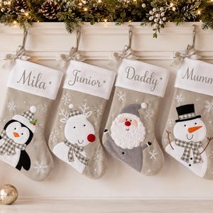 May include: Four beige Christmas stockings with white cuffs, each featuring a unique embroidered character: a penguin, a reindeer, Santa Claus, and a snowman. The cuffs are personalized with the names Mila, Junior, Daddy, and Mummy.