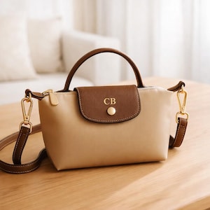 May include: A beige handbag with a brown flap and handle. The bag has a gold zipper and hardware, with the initials "CB" on the flap. A brown shoulder strap is attached with gold clasps. The bag is sitting on a wooden surface.