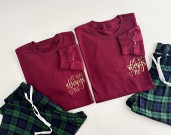 May include: Two maroon long-sleeved shirts with the text "It was always you" in gold lettering on the left sleeve. One shirt has a heart symbol before the word "you". The shirts are laid out on a white surface with a pair of green and black plaid pajama pants.