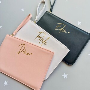 Peut inclure: Trois pochettes avec des lettres dor&eacute;es. Les pochettes sont rose, blanche et noire. La pochette rose dit "Dina" avec un c&oelig;ur. La pochette blanche dit "Bride" avec deux c&oelig;urs. La pochette noire dit "Eden" avec un c&oelig;ur.
