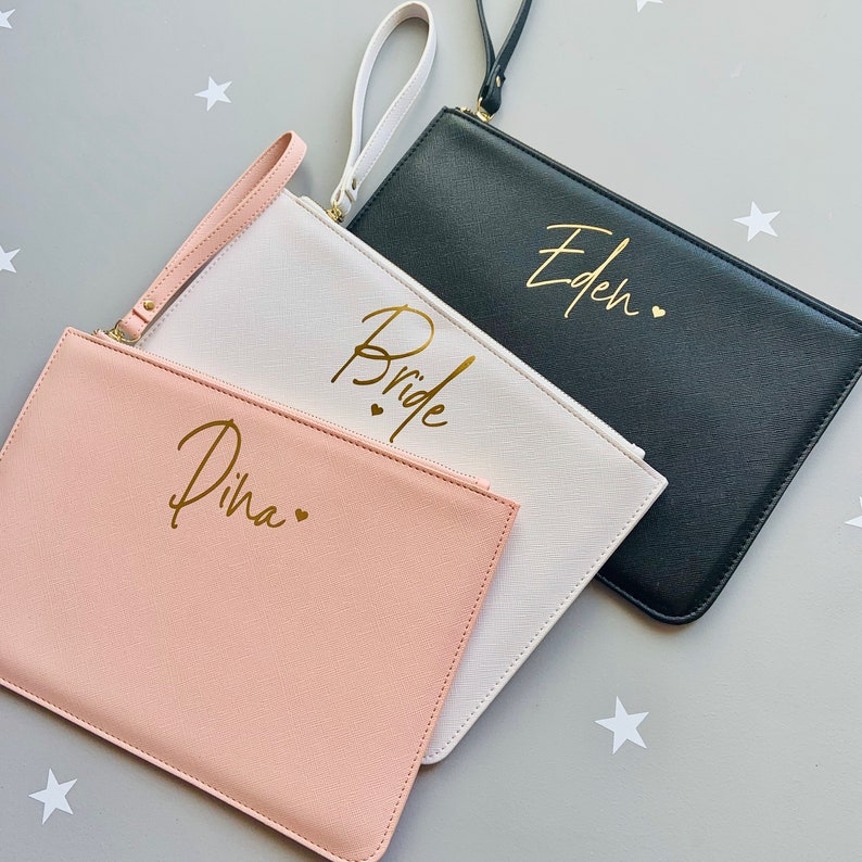 Peut inclure: Trois pochettes avec des lettres dor&eacute;es. Les pochettes sont rose, blanche et noire. La pochette rose dit "Dina" avec un c&oelig;ur. La pochette blanche dit "Bride" avec deux c&oelig;urs. La pochette noire dit "Eden" avec un c&oelig;ur.