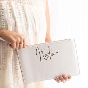 May include: A white clutch bag with a zipper closure and the name "Nadia" written in gold script with a small heart symbol.