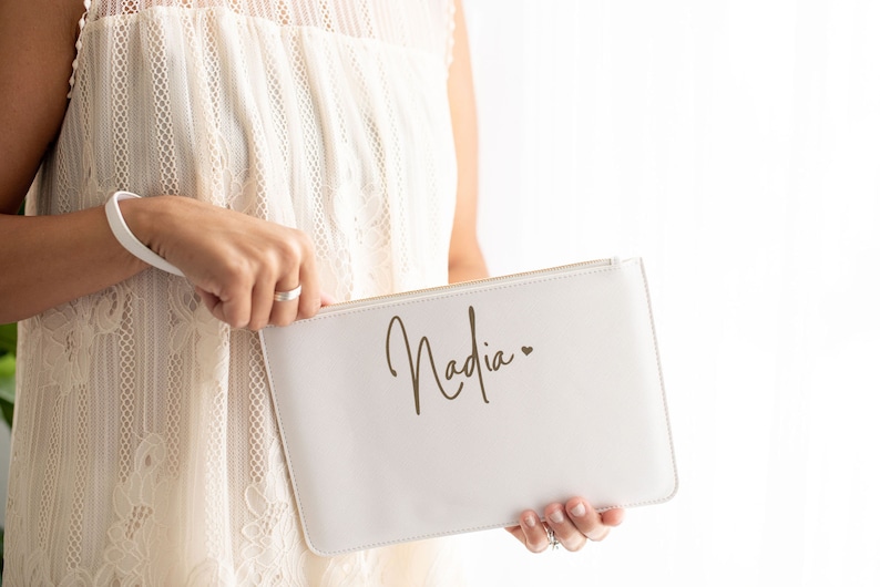 May include: A white clutch bag with a zipper closure and the name "Nadia" written in gold script with a small heart symbol.