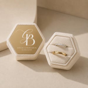 Op de afbeelding: Een zeshoekig beige fluwelen ringdoosje met een gouden deksel voorzien van het witte monogram GB en de tekst GRACE & BENJAMIN 12.08.2025. Daarnaast staat een open zeshoekig ringdoosje met twee ringen: een zilveren band met steentjes en een gouden band.