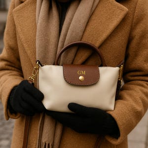 May include: A cream-coloured handbag with a brown leather flap and handle. The flap is secured with a gold-coloured button and features the initials "GM". The bag has a gold-coloured chain strap.