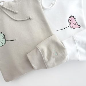 May include: Two hooded sweatshirts with embroidered dinosaurs holding phones. One sweatshirt is beige and the other is white.