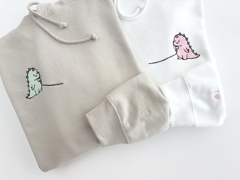 May include: Two hooded sweatshirts with embroidered dinosaurs holding phones. One sweatshirt is beige and the other is white.