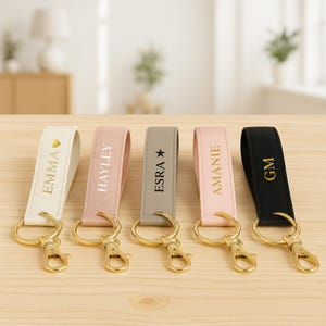 Peut inclure: Cinq porte-clés personnalisés en crème, rose, gris et noir. Chaque porte-clés est doté d'un fermoir et d'un anneau dorés, avec des noms ou initiales imprimés en or. Les porte-clés sont disposés sur une surface en bois clair.