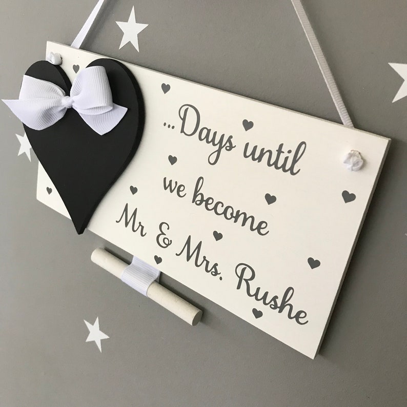 May include: White wooden sign with a black heart and a white ribbon. The sign says "...Days until we become Mr & Mrs. Rushe" with small hearts scattered around the text.