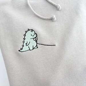 May include: A light gray hoodie with a small embroidered design of a green dinosaur holding a phone. The dinosaur is outlined in black and has a small smile.