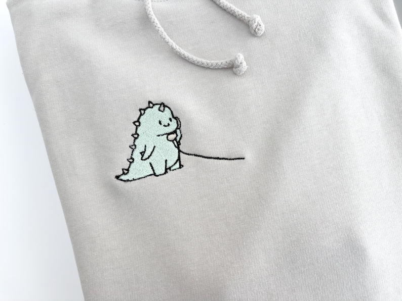May include: A light gray hoodie with a small embroidered design of a green dinosaur holding a phone. The dinosaur is outlined in black and has a small smile.