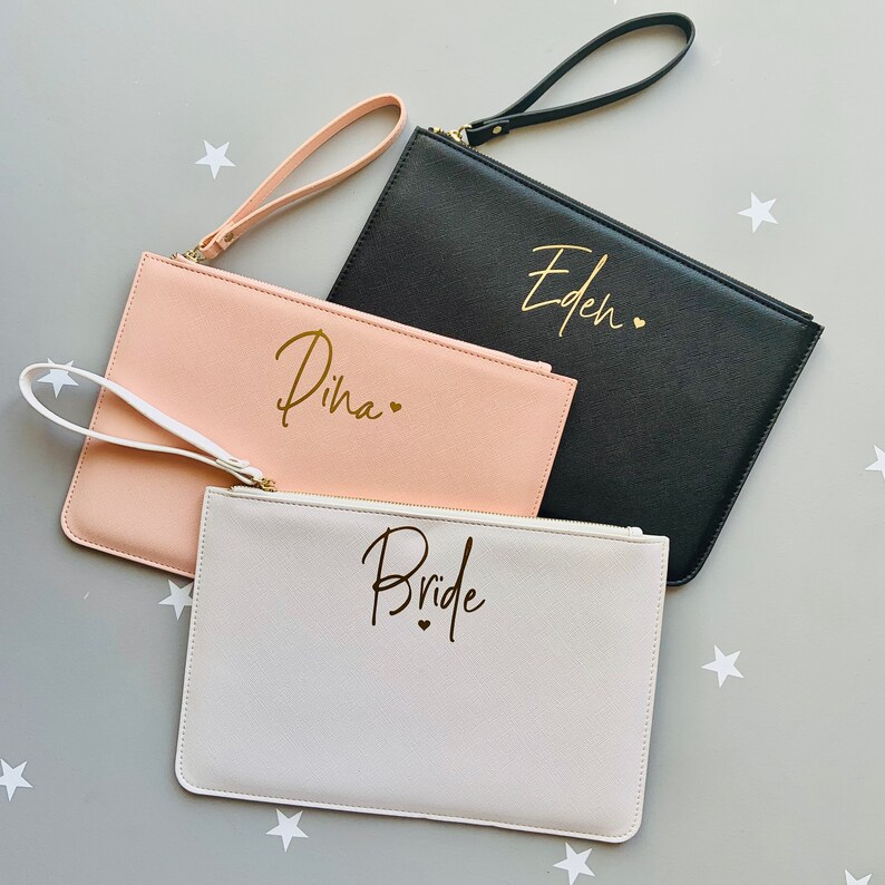 Peut inclure: Trois pochettes avec des lettres dor&eacute;es. Les pochettes sont blanches, roses et noires. La pochette blanche dit "Bride", la pochette rose dit "Dina" et la pochette noire dit "Eden".
