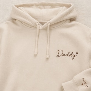 Pode incluir: Um moletom creme com capuz e cord&atilde;o. A palavra "Daddy" &eacute; bordada em escrita marrom no peito, com um pequeno cora&ccedil;&atilde;o. A manga tem o nome "ROMAN" bordado em marrom, com um cora&ccedil;&atilde;o.