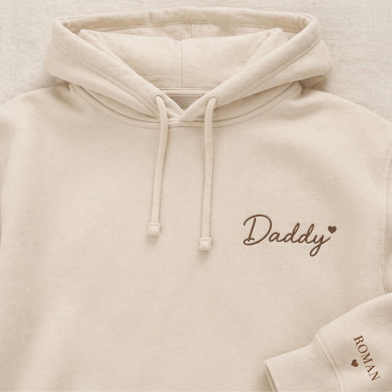 Pode incluir: Um moletom creme com capuz e cord&atilde;o. A palavra "Daddy" &eacute; bordada em escrita marrom no peito, com um pequeno cora&ccedil;&atilde;o. A manga tem o nome "ROMAN" bordado em marrom, com um cora&ccedil;&atilde;o.