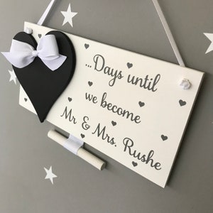 May include: White wooden sign with a black heart and a white ribbon. The sign says "...Days until we become Mr & Mrs. Rushe" with small hearts scattered around the text.