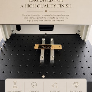 May include: A laser engraving machine with a gold-colored rectangular tag featuring the name ADRIANA centered on its black work surface. The machine displays text about high-quality finishes, precision engraving, and permanent results.