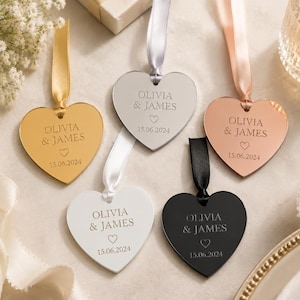 May include: Five heart-shaped ornaments in gold, silver, rose gold, white, and black finishes. Each ornament features a ribbon loop and is engraved with the text OLIVIA & JAMES, a small heart icon, and the date 15.06.2024 in a centered layout.