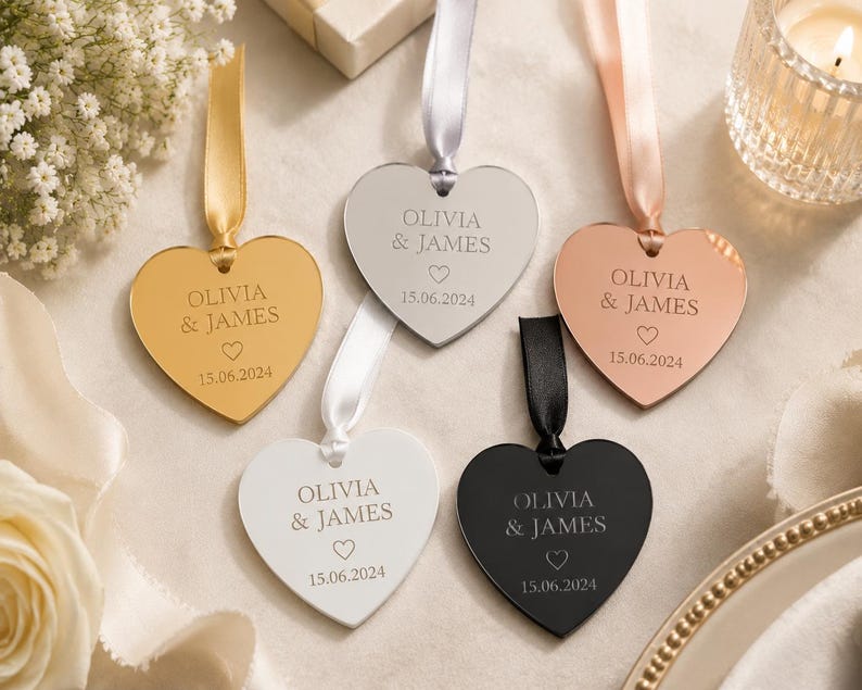 May include: Five heart-shaped ornaments in gold, silver, rose gold, white, and black finishes. Each ornament features a ribbon loop and is engraved with the text OLIVIA & JAMES, a small heart icon, and the date 15.06.2024 in a centered layout.