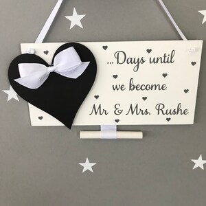 May include: White wooden sign with a black heart and a white bow. The sign says "...Days until we become Mr & Mrs. Rushe" with small hearts around the text. A white chalk board is attached to the sign.
