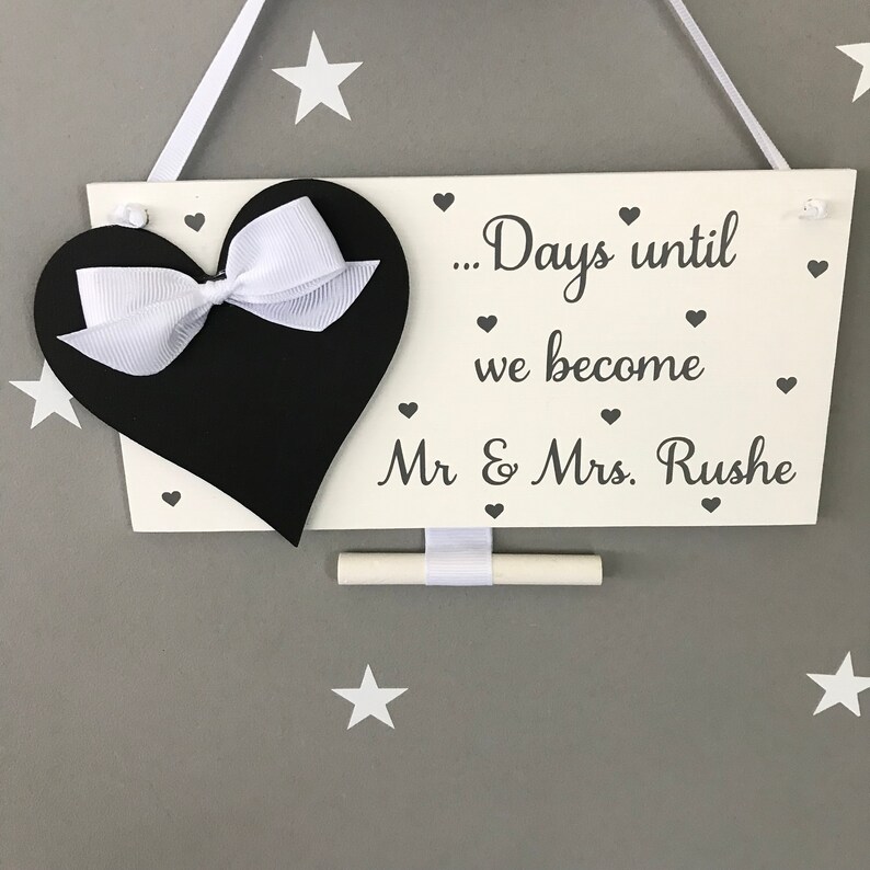 May include: White wooden sign with a black heart and a white bow. The sign says "...Days until we become Mr & Mrs. Rushe" with small hearts around the text. A white chalk board is attached to the sign.