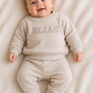 Könnte beinhalten: Ein beigefarbenes Set aus einem langärmeligen Sweatshirt und einer passenden Hose, wobei der Name ELIAS in dunkelbeigem Garn auf die Brust gestickt ist. Das Sweatshirt hat einen Rundhalsausschnitt mit Dreiecksnaht sowie gerippte Bündchen.