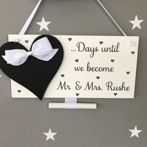 May include: White wooden sign with a black heart and a white bow. The sign says "...Days until we become Mr & Mrs. Rushe".