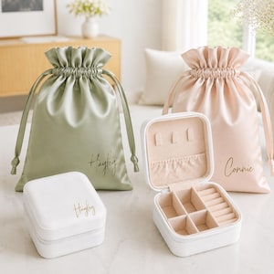 Puede incluir: Dos bolsas de satén con cordón en verde salvia y rosa pálido, personalizadas con nombres en escritura dorada. Dos joyeros cuadrados blancos las acompañan, uno cerrado y otro abierto, mostrando un interior de terciopelo beige con compartimentos.