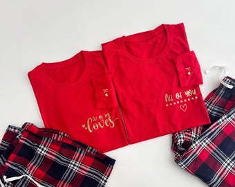 May include: Two red t-shirts with gold text that reads "All of me loves..." and "All of you". The shirts have a pocket with a gold initial and heart. Plaid pajama pants in red, navy, and white complete the set.