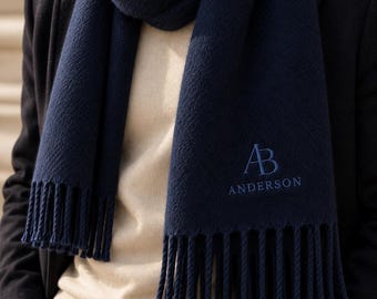 May include: A navy blue scarf featuring a textured knit pattern and fringed ends. The scarf is embroidered with the monogram AB and the name ANDERSON in a matching blue thread. The scarf is worn over a cream-colored sweater and a dark coat.