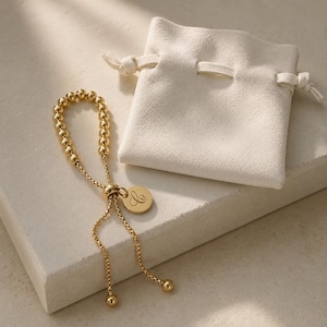 Puede incluir: Una pulsera ajustable de tono dorado que presenta una serie de pequeñas cuentas metálicas y una cadena fina. La pulsera incluye un dije circular grabado con una letra cursiva y un mecanismo deslizante para ajustar el tamaño, junto a una bolsita.